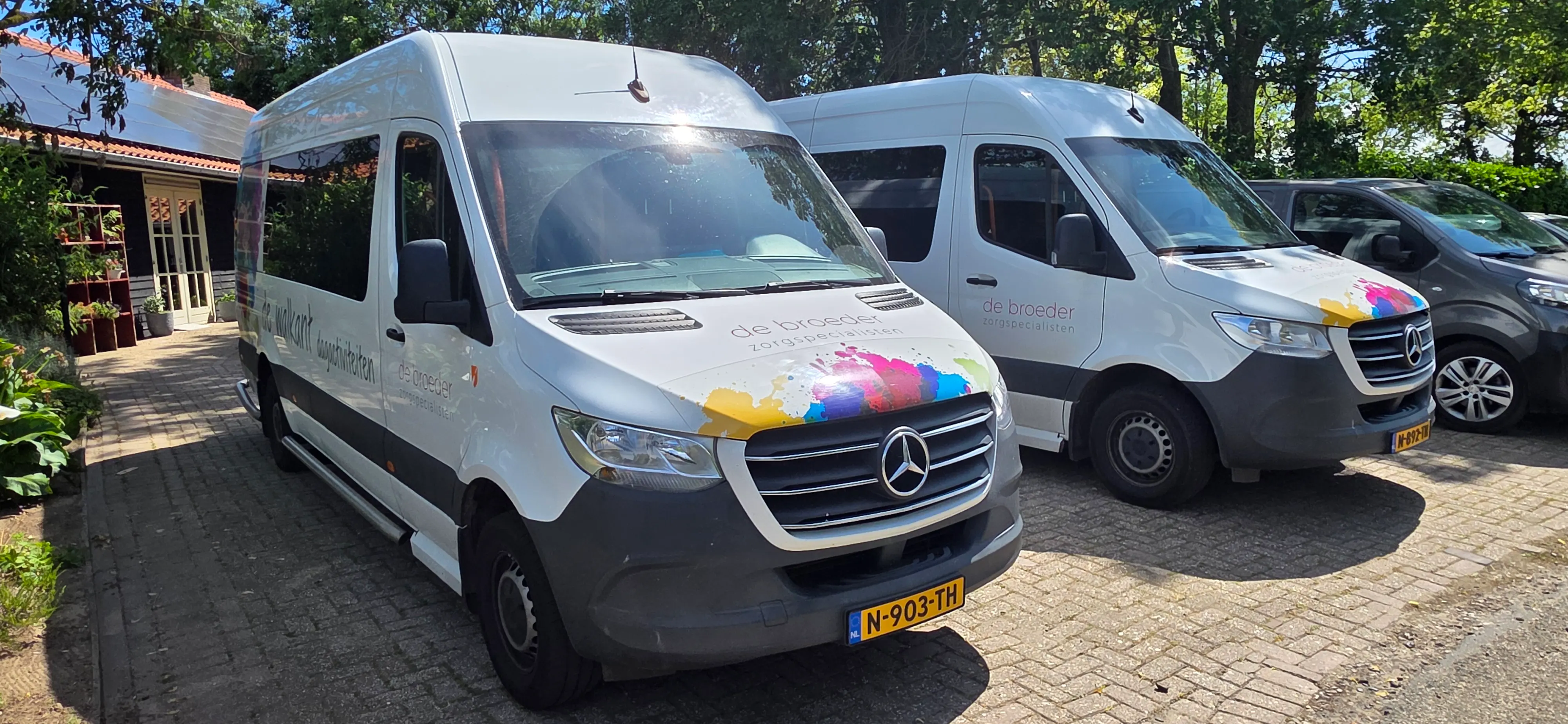Twee witte busjes van De Broeder met het kleurrijke spetterslogo, klaar voor het vervoer van en naar de dagbesteding.