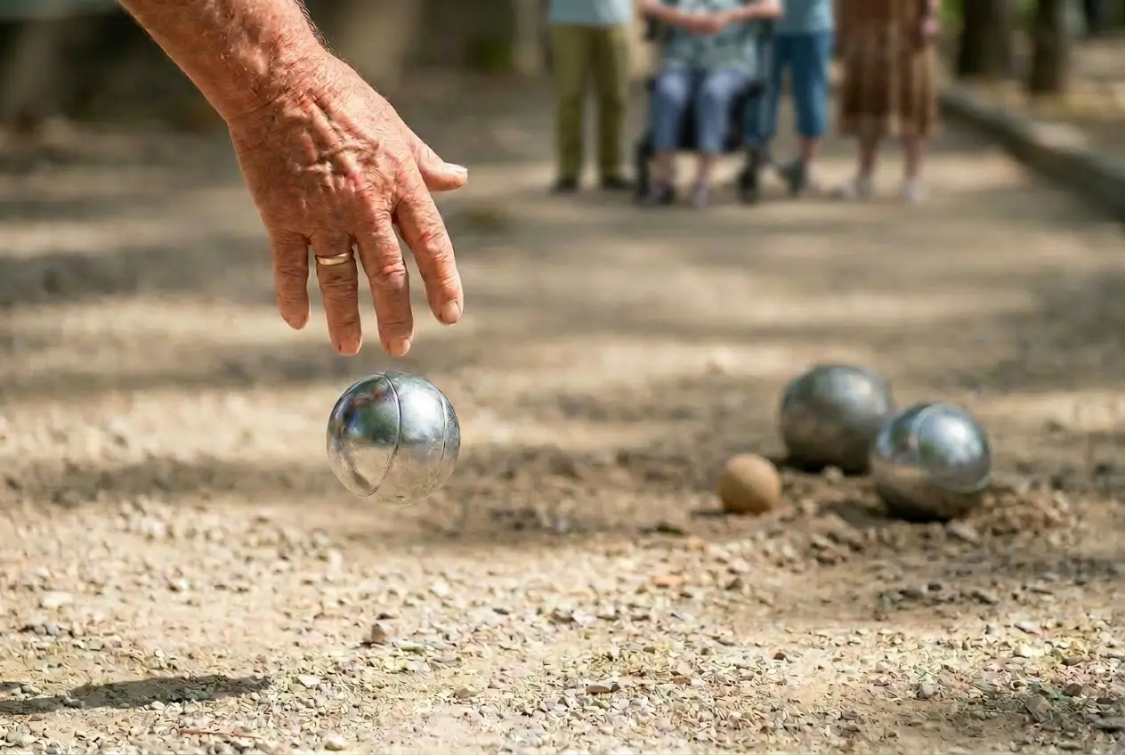 Een oudere hand werpt een zilveren jeu de boules-bal op een gravelbaan bij ActivityPlus, met wat wazige figuren op afstand.