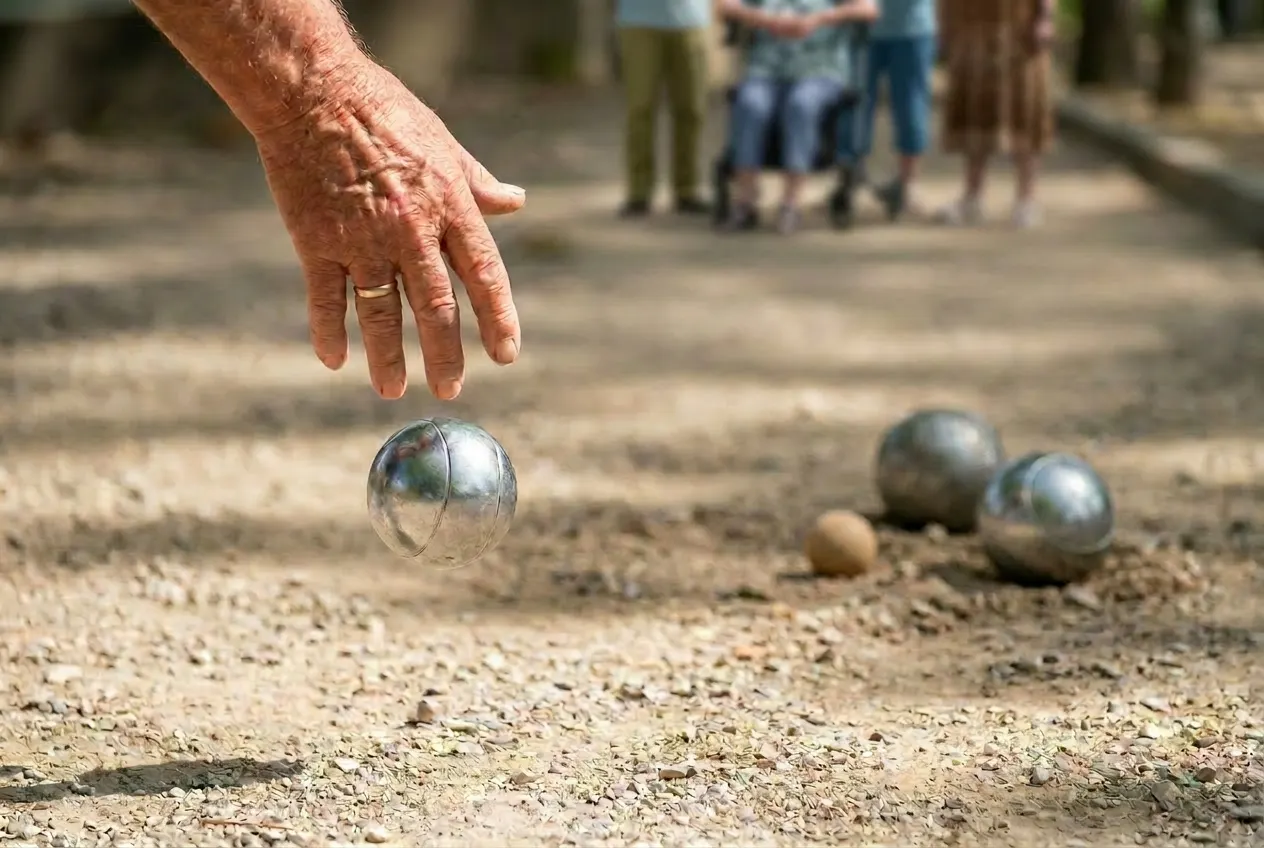 Een oudere hand werpt een zilveren jeu de boules-bal op een gravelbaan, met wat wazige figuren op afstand.