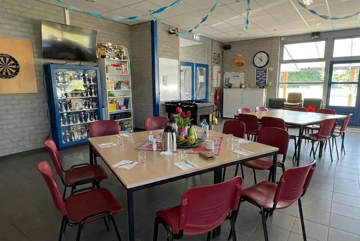 De gezellig ingerichte kantine van ActivityPlus: gedekte tafels met rode stoelen, een dartbord, een prijzenkast en tulpen op tafel.