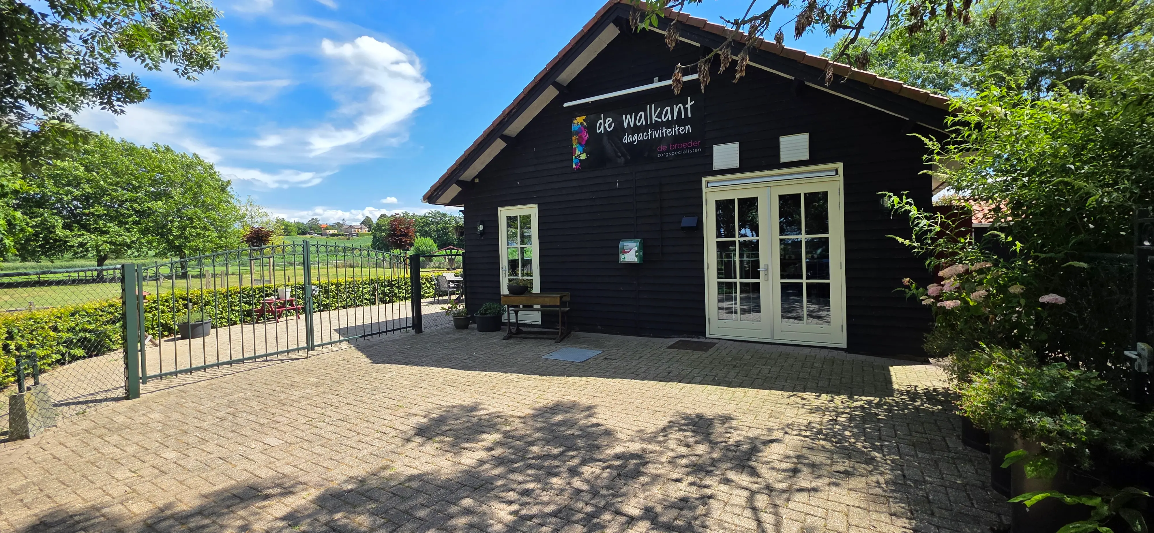De voorgevel van De Walkant: een donker houten gebouw met het bord 'de walkant dagactiviteiten' en lichtgroene openslaande deuren.